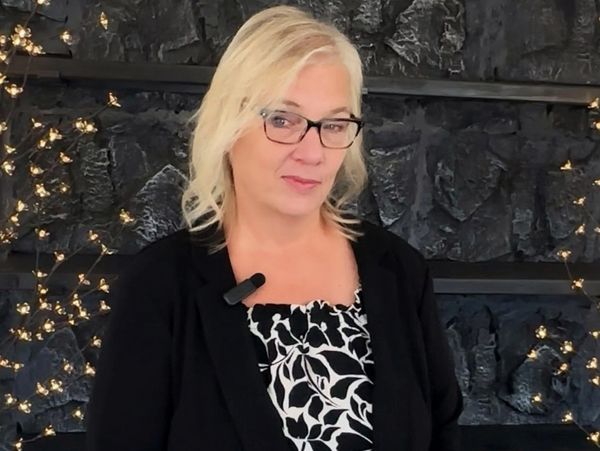 Blonde woman in glasses stands against a dark textured wall with soft lights.