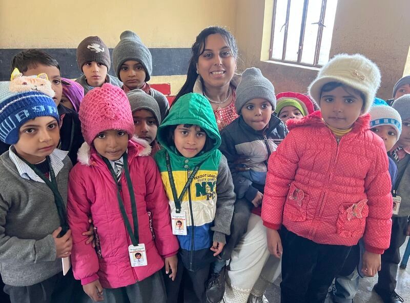 A group of children in winter clothes with a smiling woman indoors.