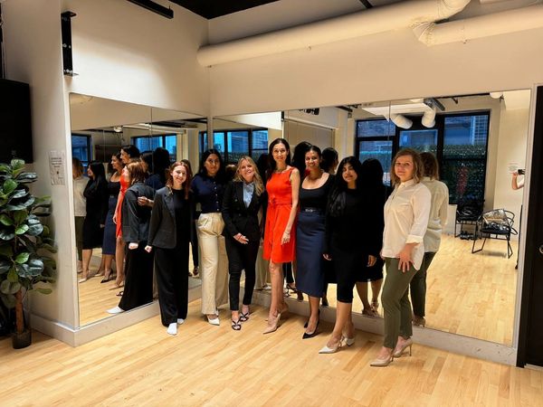 A group of eight women dressed stylishly posing in a room with large mirrors.