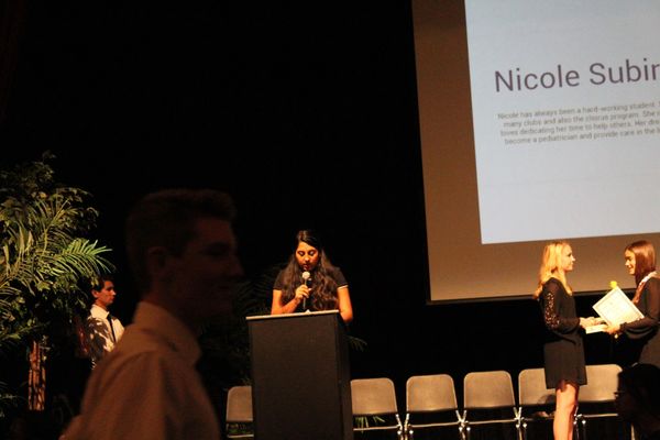 A woman speaks at a podium during an award ceremony with people receiving certificates.