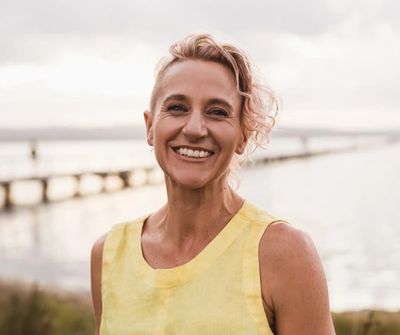 Smiling woman with short curly hair wearing a yellow dress near a waterfront.