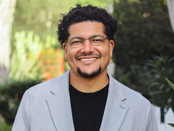 Smiling man with curly hair and glasses wearing a light gray blazer outdoors.