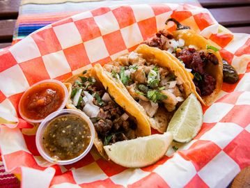 Three tacos with lime wedges and two types of salsa on a checkered paper.
