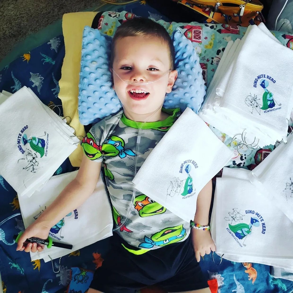 Smiling boy with nasal oxygen lying on bed surrounded by Dino-Mite Hero cloths.