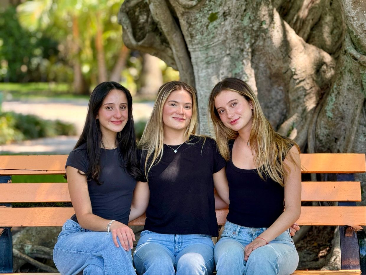 Three young women in black tops and jeans sitting on an orange bench outdoors.