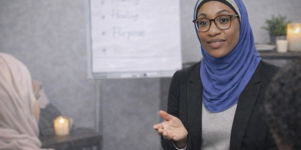 A woman in a hijab and glasses speaking in a group setting with a flip chart in the background.