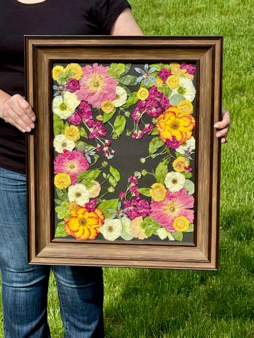 Person holding a framed floral artwork with vibrant pressed flowers on black background.