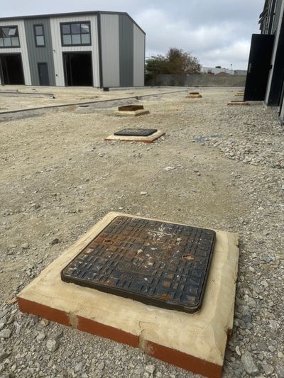Manhole covers set in brick bases on a gravel surface near industrial buildings.