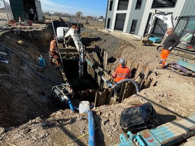 Construction workers operate excavators in a large trench near a commercial building.