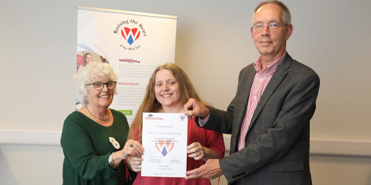 Three people smiling, holding a scholarship certificate together indoors.