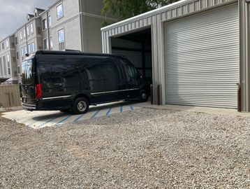 A black van parked partially in a garage with gravel and buildings nearby.