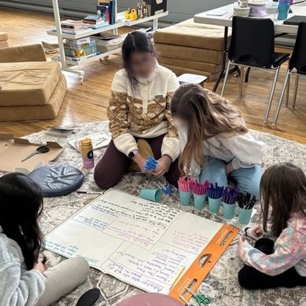 Four children collaborate on a large poster while sitting on a carpet in a cozy room.