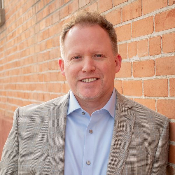 Man in a light blue shirt and checkered blazer smiling against a brick wall.