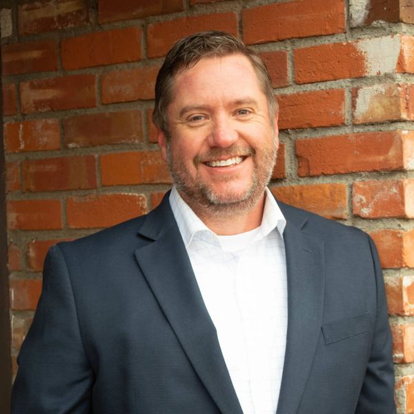 Smiling man in a navy blazer and white shirt stands against a brick wall.