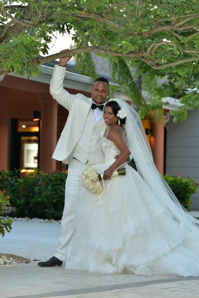 Smiling bride and groom in white attire stand under a leafy tree outdoors.