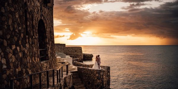 Couple standing on a stone balcony overlooking the ocean at sunset.