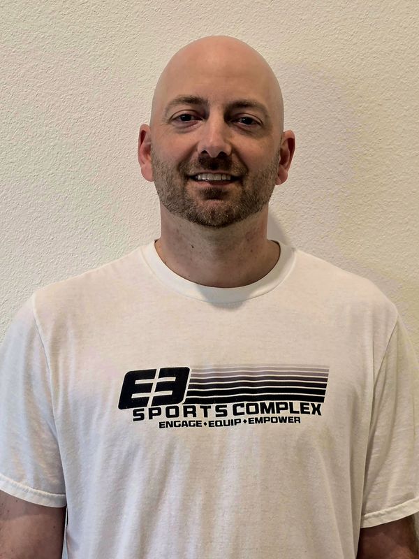 Smiling bald man wearing a white E3 Sports Complex t-shirt.