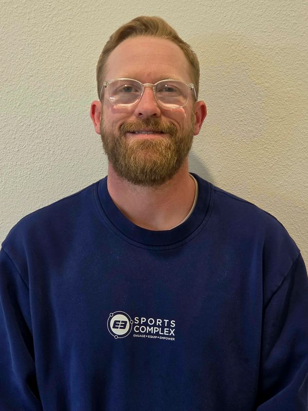 A smiling man with glasses and a beard wearing a navy blue Sports Complex sweatshirt.