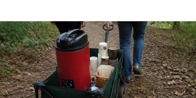 Two people pull a wagon with a red beverage dispenser and cups on a forest path.