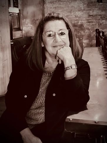 A woman with glasses sitting at a table, smiling gently.