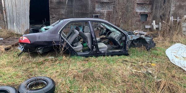 Stripped car frame without doors and front parts in a grassy yard near a barn.