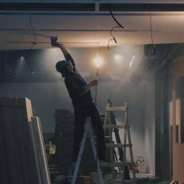 Worker installing ceiling fixtures using a ladder at night.