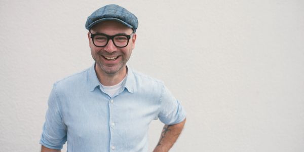 Smiling man in glasses and cap stands against a plain wall.