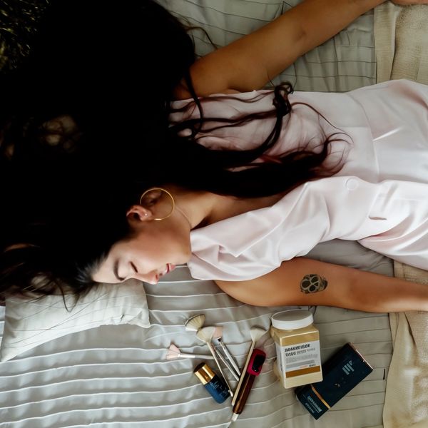 Woman lying on a bed surrounded by makeup brushes and cosmetics.