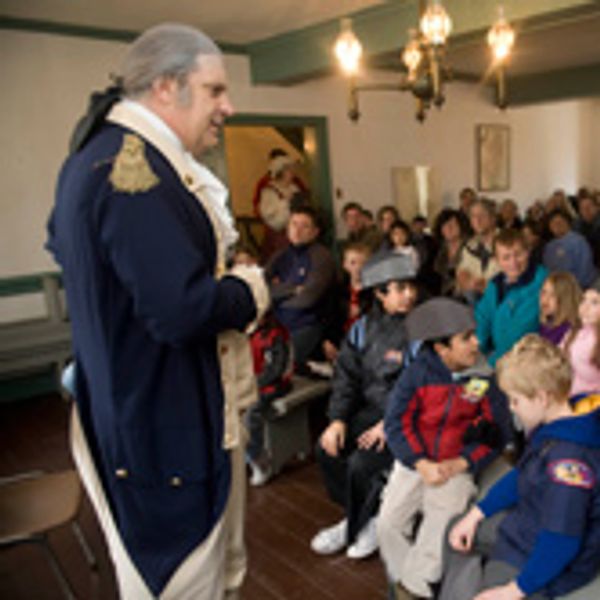 A man in historical costume speaks to a group of children and adults in a room.