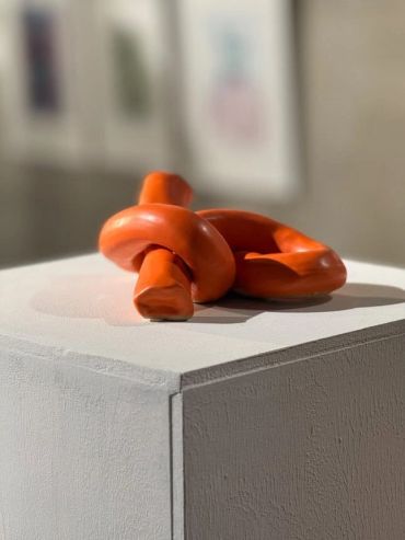 Abstract orange sculpture with twisted loops on a white pedestal in a gallery.