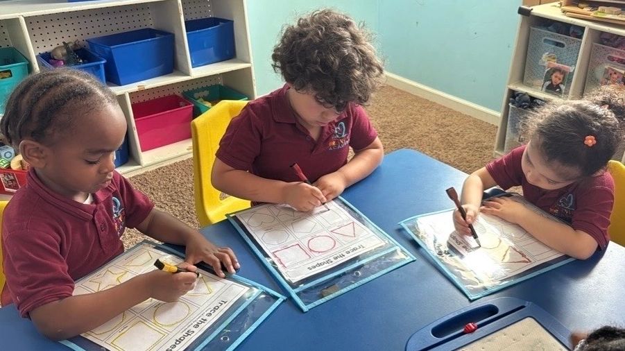 Three children practicing shapes with markers at a blue table in a classroom.