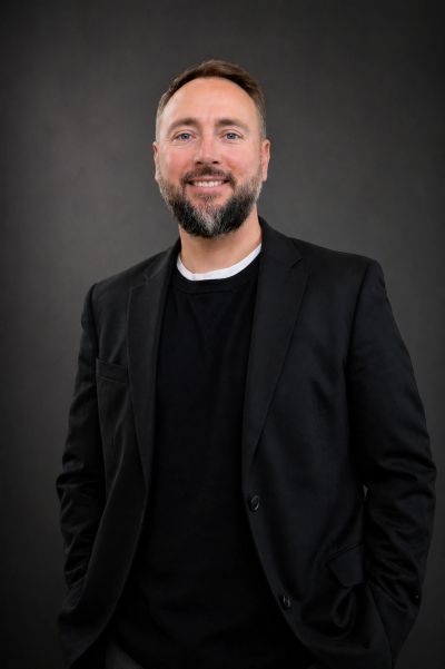 Smiling man in black blazer with hands in pockets against dark background.