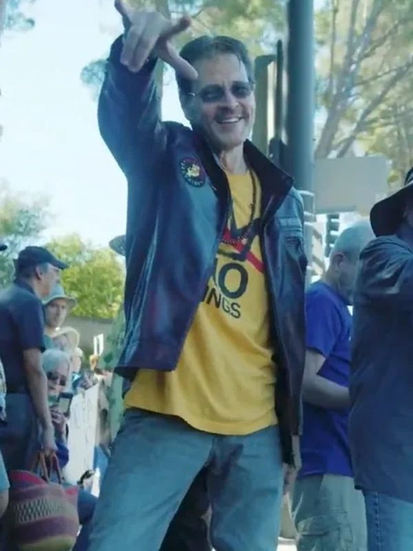 Man in leather jacket and yellow shirt smiling and making a hand gesture outdoors.