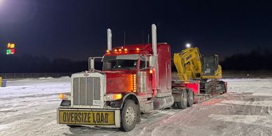 Red semi-truck carrying heavy construction equipment labeled 'Oversize Load' in snowy night.