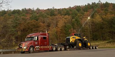 A red truck hauling heavy machinery on a trailer against a forest backdrop.