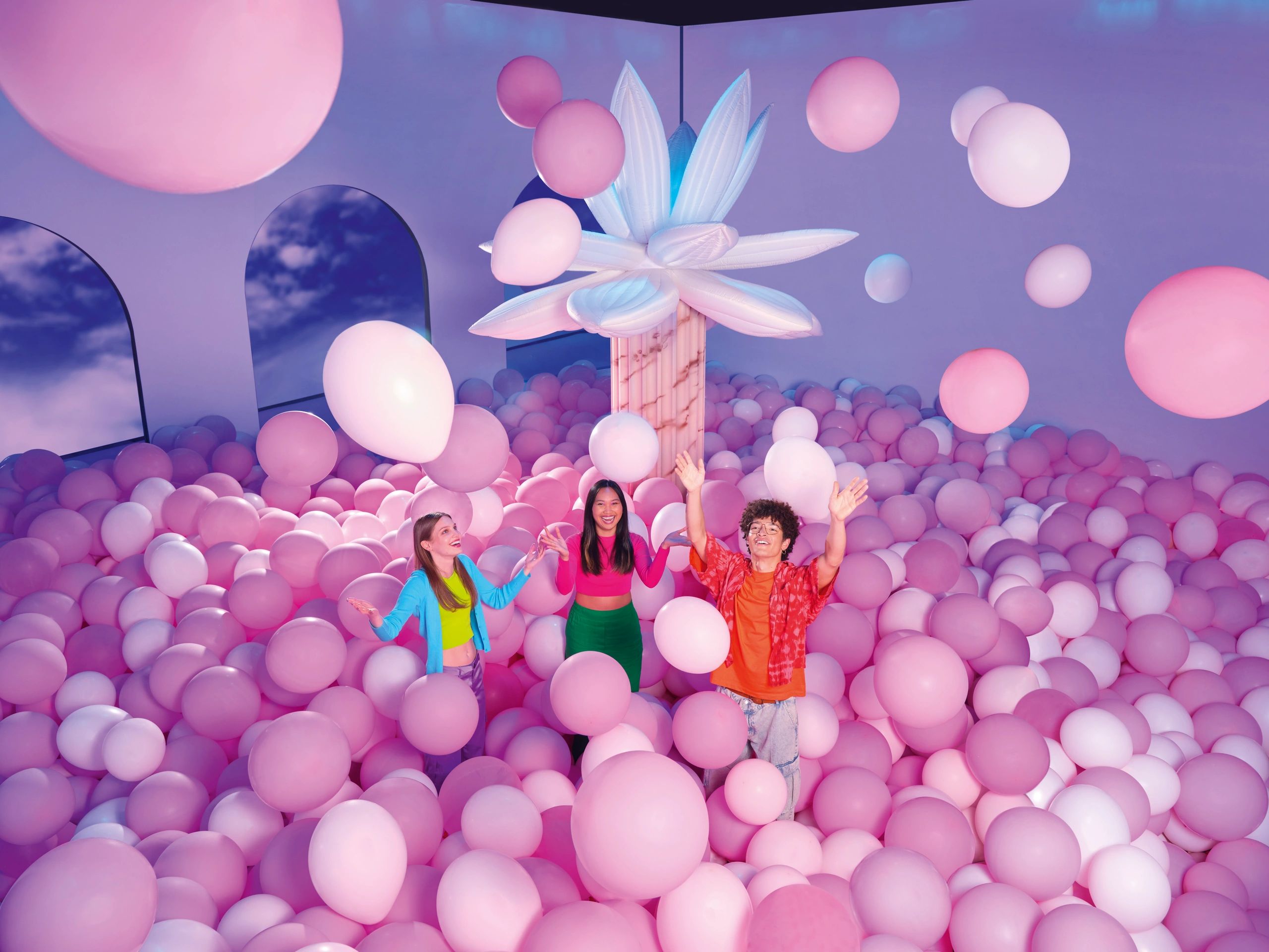Three joyful people surrounded by pink and white balloons in a vibrant room.