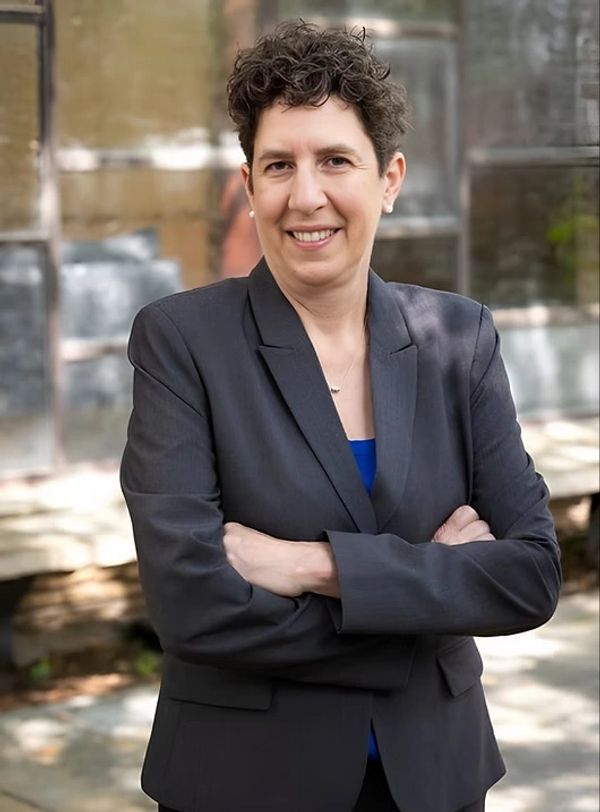 Smiling woman in a dark blazer with arms crossed outdoors.