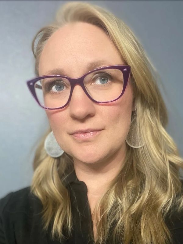 Headshot of a blonde woman with blue eyes, purple glasses, large silver earrings, and a black shirt