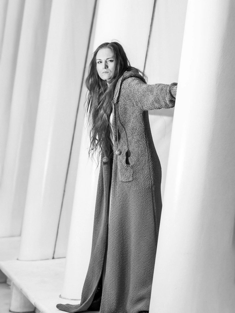 Woman in a long coat among white pillars, looking serious.