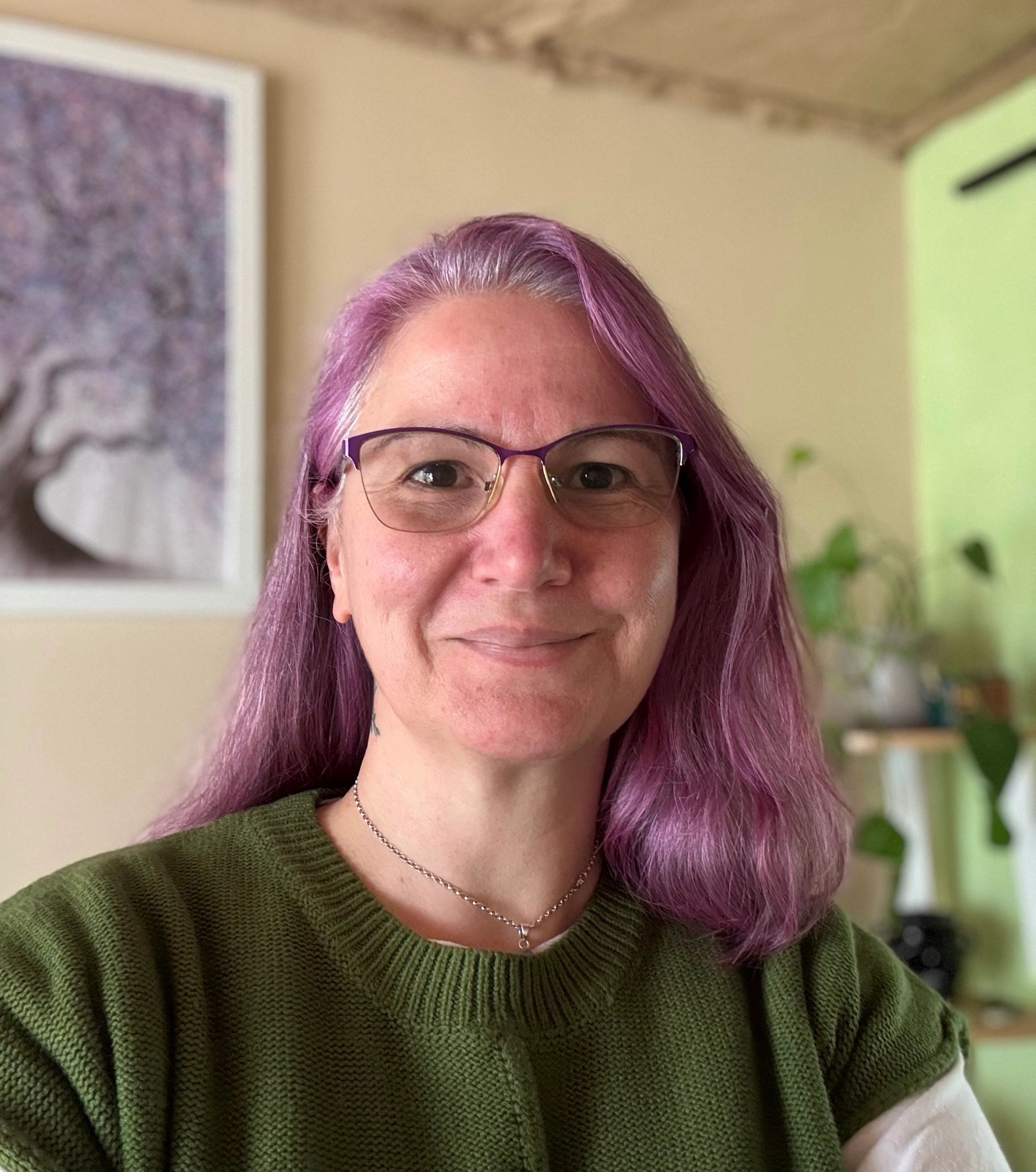 A woman with soft lavender-purple hair and matching glasses, smiling gently in a calm setting. 