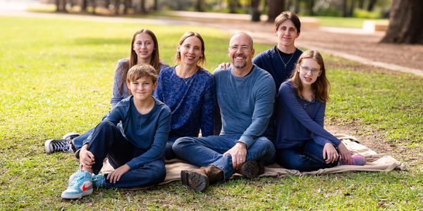 A smiling family of six sitting on a blanket in a sunny park.