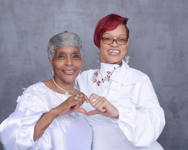 Two women in white clothing forming a heart with their hands.