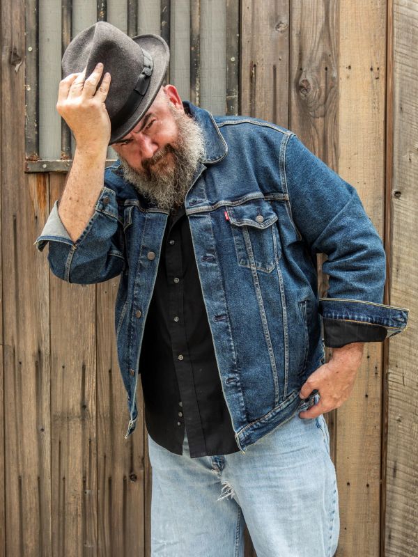 Man tipping his hat wearing a denim jacket and light jeans against a wooden fence.