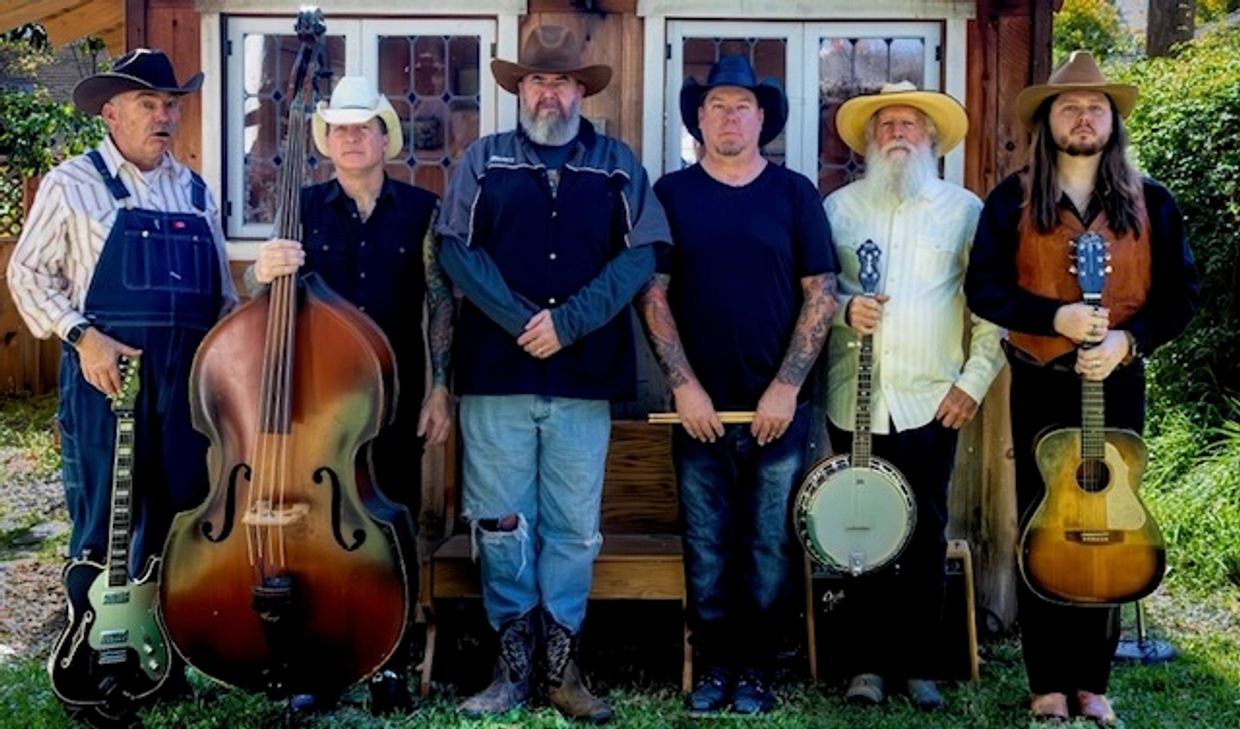 Six men in cowboy hats holding musical instruments outside a rustic building.
