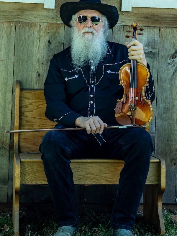 Elderly man with a long white beard holding a violin and bow, seated on a wooden bench.