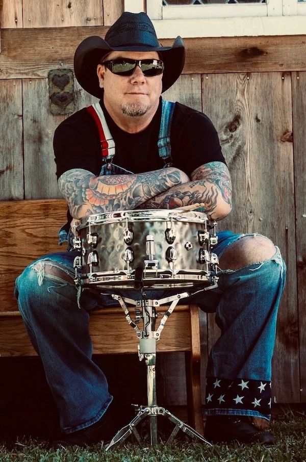 Tattooed man in cowboy hat with a snare drum sits against rustic wooden wall.