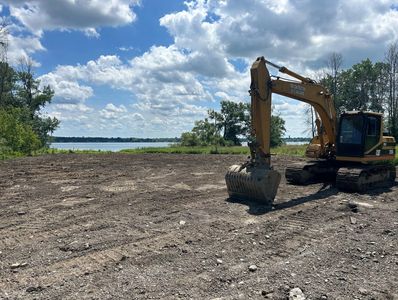 Excavator on cleared land near a lake under a partly cloudy sky.