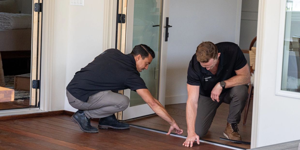Two men inspecting a wooden floor near a glass door inside a modern home.