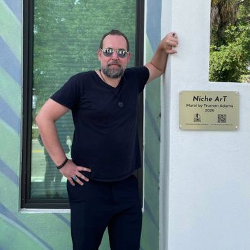 Man in black standing by a plaque for a mural by Truman Adams.