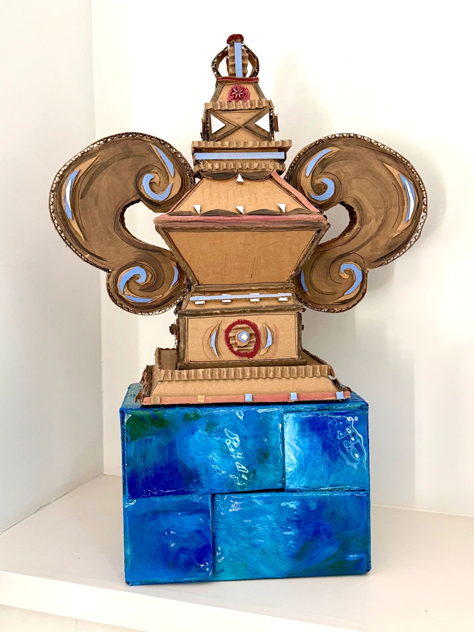 Cardboard sculpture with intricate designs, mounted on a blue painted block.
